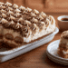 Easy Eggless Tiramisu - Thefoodiebunch Eggless Tiramisu With Piped Cream Rosettes And Cocoa Dusting In White Dish, Corner Slice On Plate Beside It