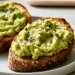 Two Slices Of Mashed Avocado Toast On Whole Grain Bread With Black Pepper And Salt On A White Plate