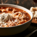 Lasagna Soup With Ground Beef, Fusilli, Ricotta Dollop, And Melted Mozzarella In A White Bowl